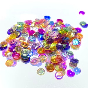 May include: A pile of iridescent sequins in various colors, including pink, purple, blue, green, and gold. The sequins are scattered on a white background.