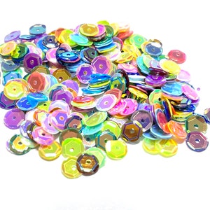 May include: A large pile of iridescent sequins in various colors, including pink, purple, blue, green, yellow, and orange. The sequins are hexagonal in shape and have a shiny, reflective finish.
