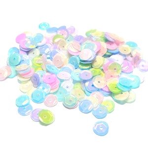 May include: A pile of iridescent sequins in pastel colors, including pink, blue, yellow, and green. The sequins are round with a hole in the center, and they are scattered on a white background. These are ideal for craft projects.