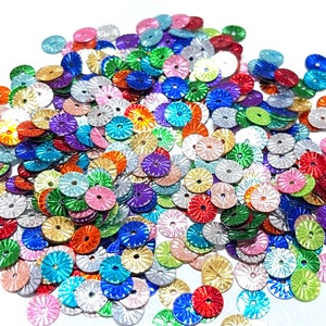 May include: A large pile of colorful, metallic, round sequins with a scalloped edge. The sequins are arranged in a random pattern and are all different colors, including gold, silver, red, blue, green, and purple.