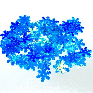 May include: A pile of iridescent blue snowflake-shaped sequins. The sequins are scattered on a white surface.