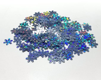 May include: A pile of iridescent silver snowflake-shaped glitter. The glitter is scattered on a white surface.