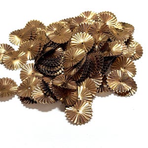 May include: A pile of small, metallic gold-colored paper rosettes. Each rosette has a circular shape with a central hole and radiating, pleated edges. The rosettes are scattered against a white background, creating a textured, decorative effect.