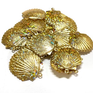 May include: A pile of gold-colored plastic seashells with iridescent glitter. The shells have a scalloped edge and a small loop at the top.