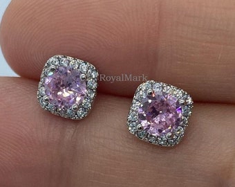 May include: A pair of square-shaped stud earrings featuring a round, light pink center stone surrounded by a halo of small, clear, sparkling stones set in a metallic frame. The text RoyalMark is visible in small, faint lettering near the earrings.