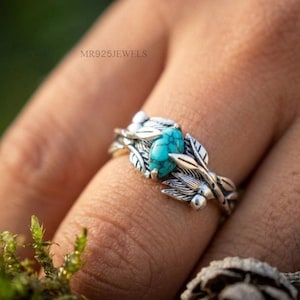 Puede incluir: Un anillo de plata con una piedra turquesa de corte marquesa engastada en una banda diseñada con hojas y enredaderas de metal detalladas. La banda tiene un acabado oxidado para resaltar la textura del follaje. El texto MR925JEWELS es visible.
