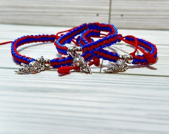Puede incluir: Tres pulseras de macramé hechas a mano en rojo y azul. Cada pulsera presenta un diseño tejido y un dije plateado. Las pulseras se exhiben sobre una superficie de madera de color claro.