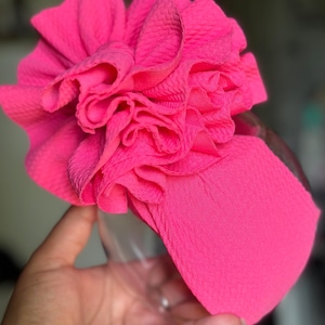 May include: A vibrant pink headband with a large, ruffled fabric flower. The headband has a textured finish and is designed to be worn around the head. The flower is the main feature, adding a decorative touch to the accessory.