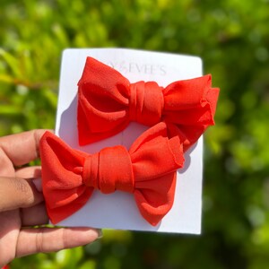 May include: Two bright orange fabric hair bows on a white card. The bows are tied in a knot in the center.