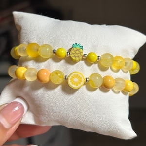 Puede incluir: Dos pulseras de cuentas amarillas con un dije de piña y limón. Las pulseras están sobre un fondo blanco.