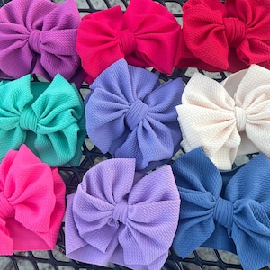 Peut inclure: Collection de bandeaux pour bébés avec de grands nœuds texturés. Les bandeaux sont disponibles dans différentes teintes, notamment rose, violet, bleu, vert et crème. Les nœuds sont en tissu doux et texturé.