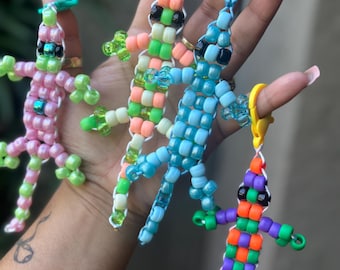 Può includere: Quattro portachiavi a forma di lucertola di perline rosa, verdi, blu e viola. Ogni lucertola ha una diversa combinazione di colori, con occhi di perline nere e una chiusura in plastica. I portachiavi sono fatti di piccole perline colorate infilate insieme.