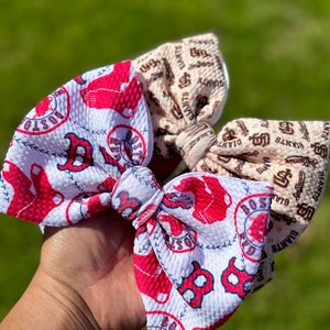 May include: Two fabric hair bows with a white background and red and brown logos. The left bow has a red logo with the text "BOSTON" and a baseball. The right bow has a brown logo with the text "GIANTS".