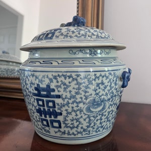 Puede incluir: Un jarrón de cerámica azul y blanco con tapa. El jarrón presenta un patrón floral y de vid con un símbolo de doble felicidad. La tapa tiene una pequeña escultura azul en la parte superior. El jarrón está sobre una superficie de madera.