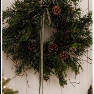 May include: A circular wreath constructed from dark green pine needles, thin brown twigs, and assorted green leaves. The wreath is decorated with several brown pinecones and small, round, textured seed pods, featuring a long, light green velvet ribbon.