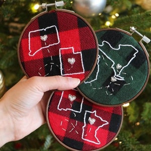 May include: Hand-held, embroidered Christmas ornaments. The ornaments feature state outlines with a heart, stitched on red and green plaid fabric. Each ornament is framed in a wooden hoop, perfect for holiday decor.