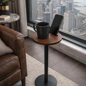 Peut inclure: Une table d'appoint moderne avec un plateau en bois foncé et une base en métal noir. Une tasse noire et un smartphone sont posés sur la table. La table est près d'une fenêtre avec vue sur la ville.