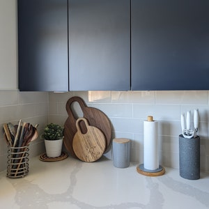 May include: A kitchen countertop with a gray and white tiled backsplash. There are two wooden cutting boards, a gray ceramic canister, a white paper towel holder, and a gray ceramic container with a black lid.