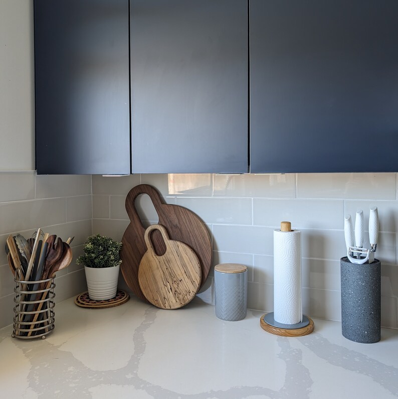 May include: A kitchen countertop with a gray and white tiled backsplash. There are two wooden cutting boards, a gray ceramic canister, a white paper towel holder, and a gray ceramic container with a black lid.