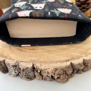 A black book sleeve featuring a pattern of white ghosts, tan, and green mushroom-like figures on a dark backdrop. The sleeve has a book inside. It rests on a wooden surface.