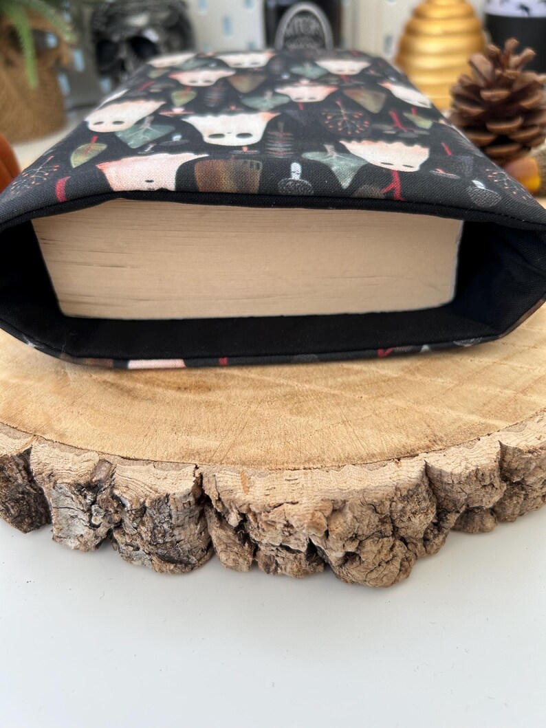 A black book sleeve featuring a pattern of white ghosts, tan, and green mushroom-like figures on a dark backdrop. The sleeve has a book inside. It rests on a wooden surface.