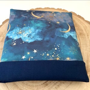 A fabric book sleeve with a celestial design and a navy blue base. The top section shows a night sky with gold crescent moons, stars, and clouds in shades of blue and teal. The lining is a solid navy blue.