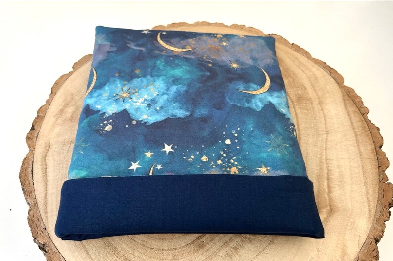 A fabric book sleeve with a celestial design and a navy blue base. The top section shows a night sky with gold crescent moons, stars, and clouds in shades of blue and teal. The lining is a solid navy blue.