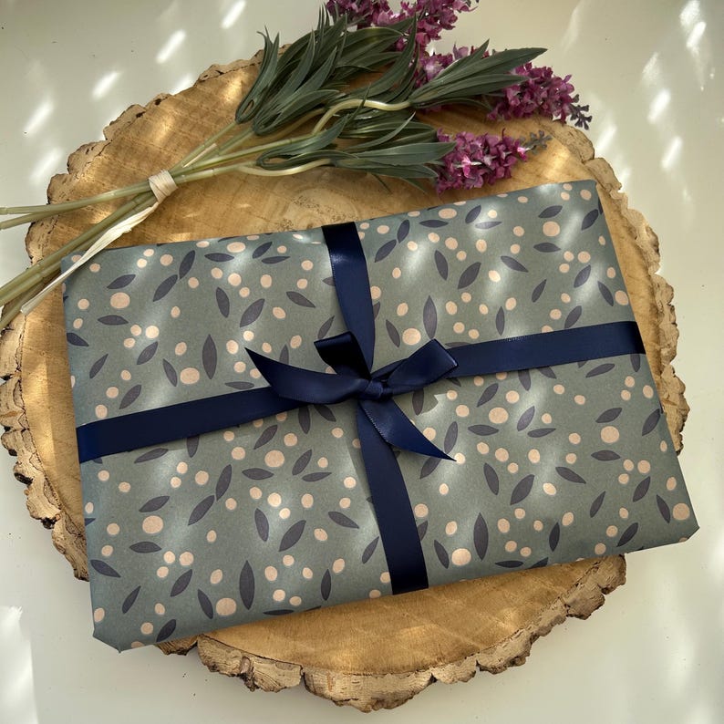 A book sleeve wrapped in blue-grey paper with a pattern of white dots and dark blue leaves, tied with a navy blue ribbon and bow. It sits on a wooden slice, with purple lavender sprigs. A decorative gift for any occasion.