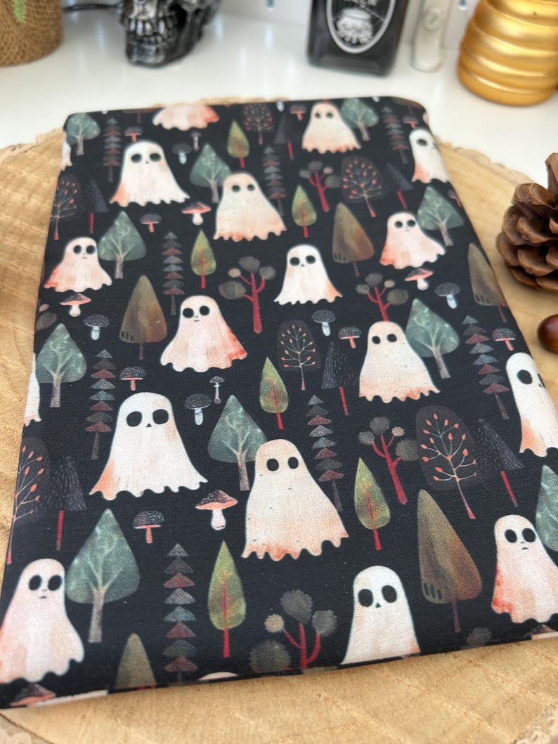 Black fabric book cosy featuring a repeating pattern of white ghosts, trees, and mushrooms. The ghosts have a simple, rounded shape with two black eyes. The trees are various shades of green and brown. The design has a spooky, Halloween theme.