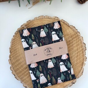 A black fabric book cosy with a repeating pattern of ghosts, trees, and mushrooms in white, peach, and green. The design is set against a dark background. The book cosy is resting on a wooden surface and has a brown paper belly band.