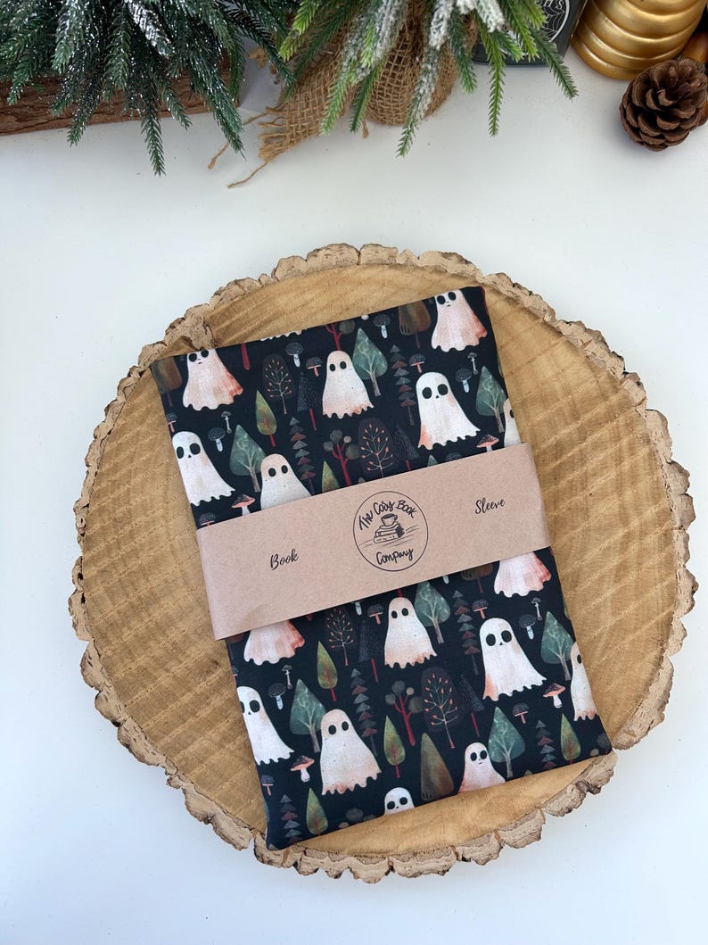 A black fabric book cosy with a repeating pattern of ghosts, trees, and mushrooms in white, peach, and green. The design is set against a dark background. The book cosy is resting on a wooden surface and has a brown paper belly band.
