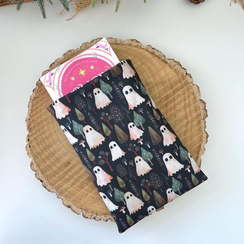 A black book sleeve with a repeating pattern of white ghosts, trees, and mushrooms. A book is partially visible. A wooden slice forms the background.