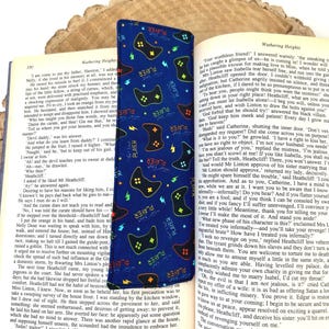 May include: A blue bookmark with a pattern of black video game controllers and the word "PLAYER" in different colours. The bookmark is inserted in a book.