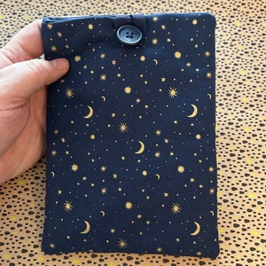 May include: A navy blue book sleeve featuring a gold celestial design of stars and crescent moons. The sleeve has a button closure. The fabric is a dark blue with a repeating pattern. Ideal for protecting books.