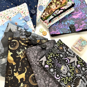 May include: A collection of fabric book covers featuring a variety of patterns, including floral, geometric, and whimsical designs. The covers are made from cotton fabric and have button fastenings. Some of the covers have a black background with gold accents.