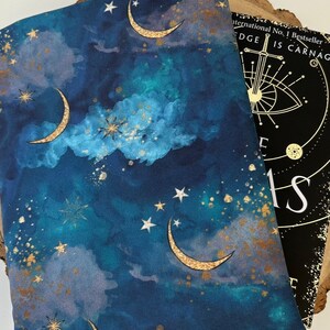 A dark blue book sleeve with a celestial design featuring gold  coloured crescent moons, stars, and clouds. The sleeve rests on a book. The book sleeve is made of fabric.