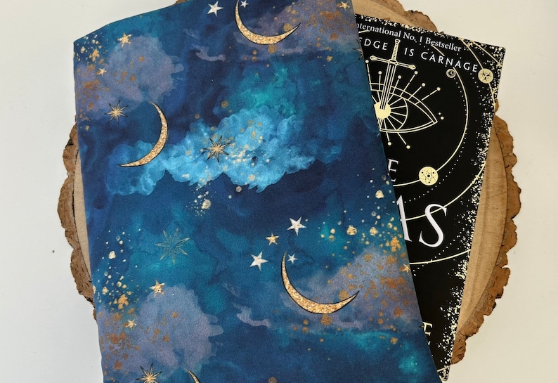 A dark blue book sleeve with a celestial design featuring gold  coloured crescent moons, stars, and clouds. The sleeve rests on a book. The book sleeve is made of fabric.