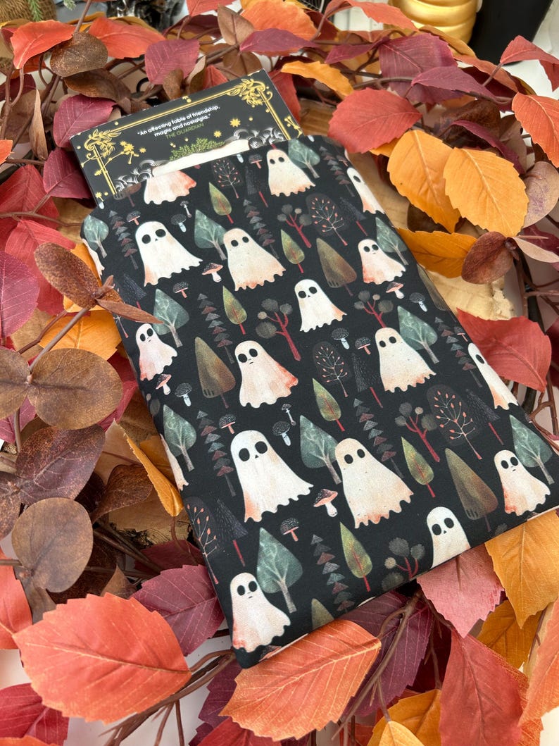 A black book sleeve with a repeating pattern of white ghosts, trees, and mushrooms. A book is partially visible. Autumn leaves form the background.
