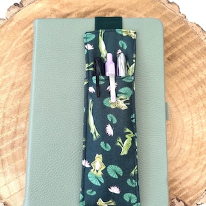 May include: A green notebook with a frog-themed pen holder attached. The pen holder is made of dark green fabric with a pattern of green frogs, pink lily pads, and green leaves. The pen holder has three pens inside.