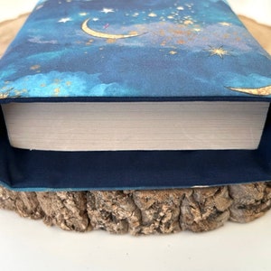 A dark blue book sleeve with a celestial design featuring gold  coloured crescent moons, stars, and clouds. The sleeve holds a book. The book sleeve is made of fabric.