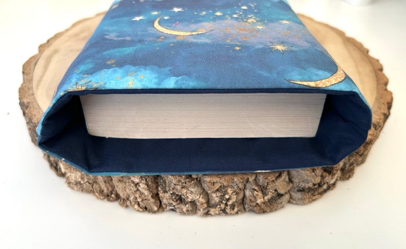 A dark blue book sleeve with a celestial design featuring gold  coloured crescent moons, stars, and clouds. The sleeve holds a book. The book sleeve is made of fabric.