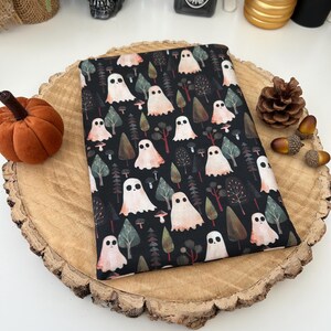 A black fabric pouch with a repeating pattern of ghosts, trees, and mushrooms. The ghosts are white with peach accents. The background is a dark, forest scene. The pouch is displayed on a wooden slice.