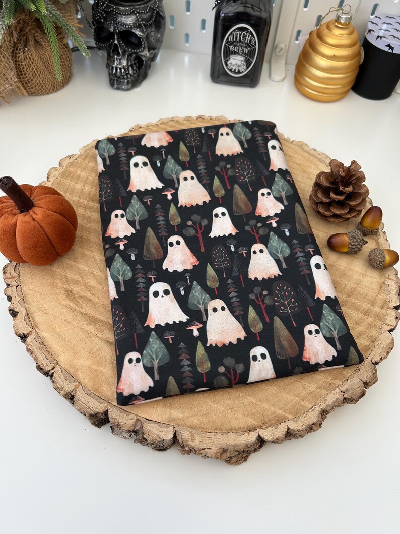 A black fabric pouch with a repeating pattern of ghosts, trees, and mushrooms. The ghosts are white with peach accents. The background is a dark, forest scene. The pouch is displayed on a wooden slice.