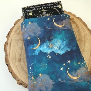 A dark blue book sleeve with a celestial design featuring gold  coloured crescent moons, stars, and clouds. The sleeve holds a book. The book sleeve is made of fabric.