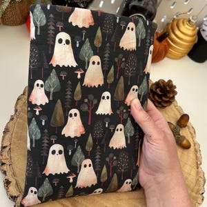 A black fabric book cosy with a repeating pattern of ghosts, trees, and mushrooms in white, peach, and green. The design is set against a dark background. The book cosy is resting on a wooden surface.