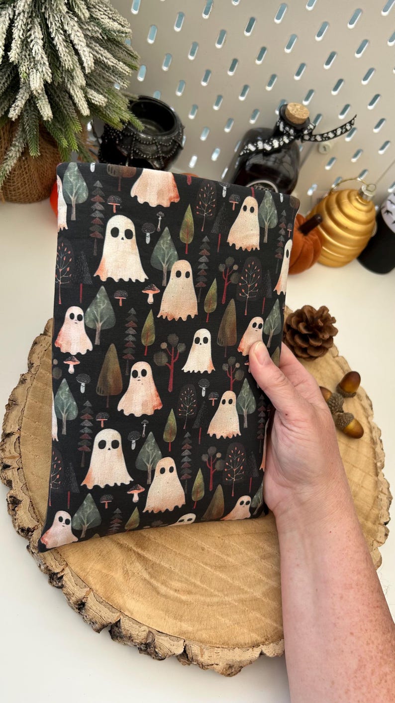 A black fabric book cosy with a repeating pattern of ghosts, trees, and mushrooms in white, peach, and green. The design is set against a dark background. The book cosy is resting on a wooden surface.