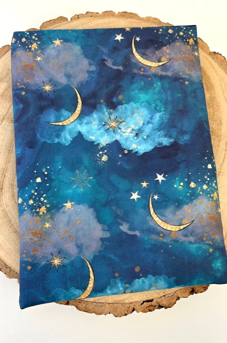 A dark blue book sleeve with a celestial design featuring gold  coloured crescent moons, stars, and clouds. The sleeve holds a book. The book sleeve is made of fabric.
