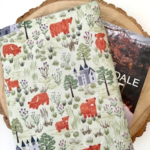 May include: A pale green fabric featuring a pattern of Highland cattle, castles, and wildflowers. A postcard with the word "DALE" is partially visible. The fabric rests on a wooden slice.