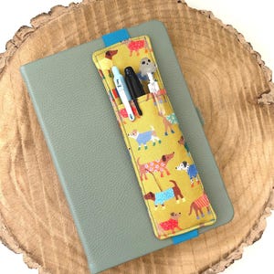 May include: A grey notebook with a yellow fabric pen holder attached to the cover. The pen holder has a pattern of dogs wearing jumpers and is secured with a blue ribbon. The pen holder contains three pens.