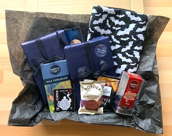 Op de afbeelding: Een zwarte cadeaudoos gevuld met verschillende artikelen, waaronder een zwarte en witte vleermuisprintstof, twee blauwe cadeaudozen, een chocolade reep, een zak koekjes, een zak koffie en een doos met chocolade roerstaafjes. De doos is bekleed met zwart tissuepapier met zilveren sterren.